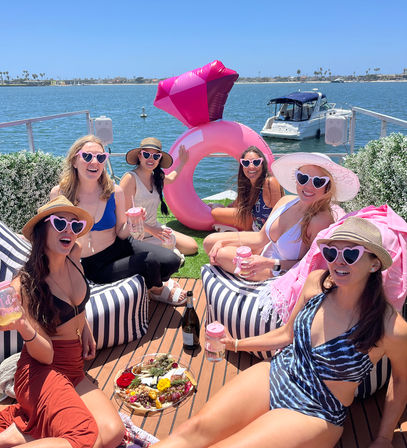 Six friends in swimsuits wearing pink heart sunglasses, sipping drinks around a charcuterie board and giant inflatable ring on a sunny marina dock by a calm bay.