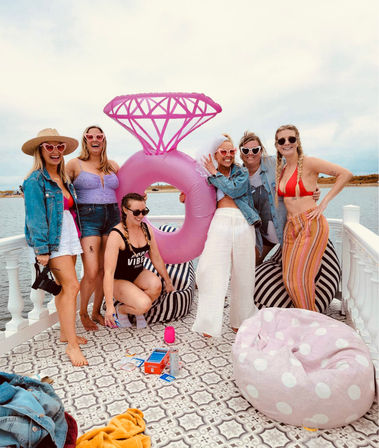 Six friends on a waterfront deck posing with a giant pink inflatable diamond ring float — summer swimwear, sunglasses, denim jackets and striped beanbags for a playful beach-party vibe