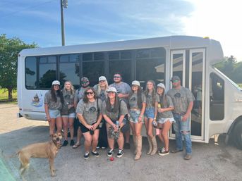 Smiling group of adults in matching camo shirts and caps posing in front of a white shuttle bus in a sunny parking lot with a brown dog in the foreground — summer group outing photo.