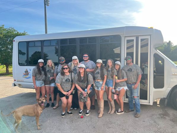 Smiling group of adults in matching camo shirts and caps posing in front of a white shuttle bus in a sunny parking lot with a brown dog in the foreground — summer group outing photo.