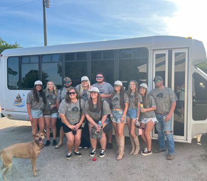 Smiling group of adults in matching camo shirts and caps posing in front of a white shuttle bus in a sunny parking lot with a brown dog in the foreground — summer group outing photo.