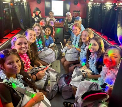 Neon-lit party bus interior with a smiling group of teenage girls wearing leis, holding backpacks and enjoying a festive ride.