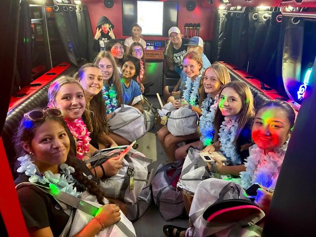 Neon-lit party bus interior with a smiling group of teenage girls wearing leis, holding backpacks and enjoying a festive ride.