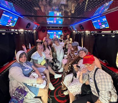 Neon-lit party bus interior with a group of adults in playful vintage/elderly-themed costumes posing on leather benches beneath a reflective diamond-plate ceiling, LED strip lights, TV screens and visible snack bags — lively group celebration.