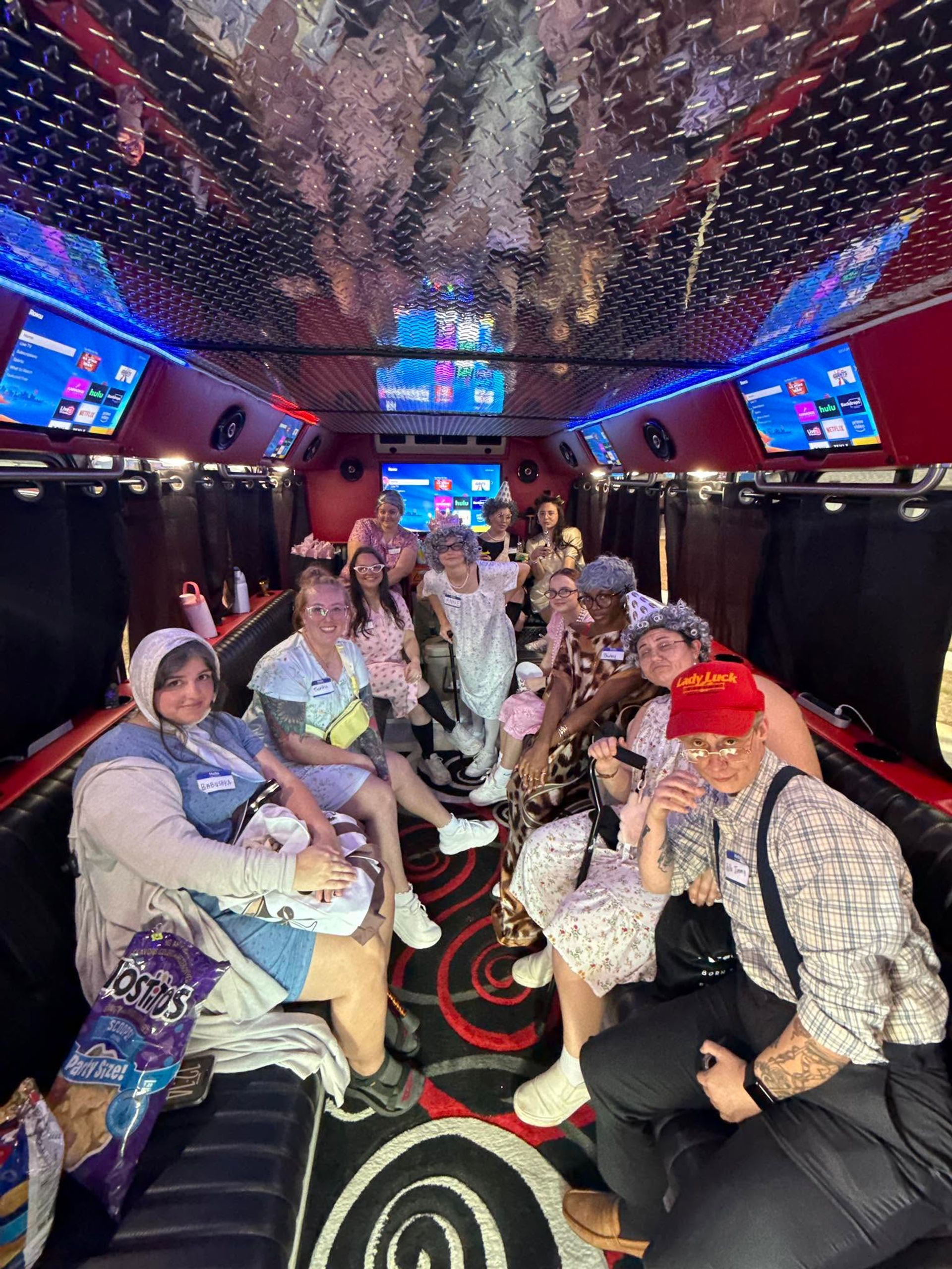 Neon-lit party bus interior with a group of adults in playful vintage/elderly-themed costumes posing on leather benches beneath a reflective diamond-plate ceiling, LED strip lights, TV screens and visible snack bags — lively group celebration.