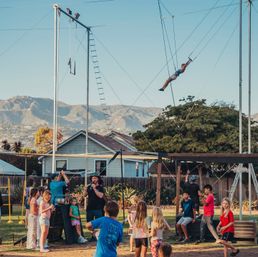 Sunlit backyard trapeze setup with an aerialist swinging high above a group of children watching, suburban houses and a mountain range in the background.