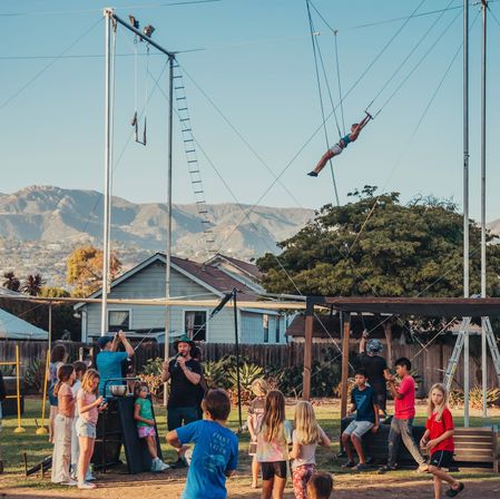 Sunlit backyard trapeze setup with an aerialist swinging high above a group of children watching, suburban houses and a mountain range in the background.