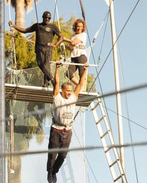 Man in a safety harness gripping a trapeze bar mid-air during an outdoor trapeze lesson, watched by two instructors on a platform with netting and ladder against a sunny blue sky.
