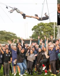 Smiling group of adults in a park raising hands on a grassy field beneath two trapeze artists mid-air holding hands during an outdoor aerial performance