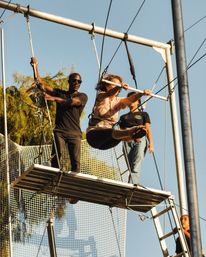 Woman takes a flying swing on an outdoor trapeze, harnessed above a safety net while instructors assist from a metal platform and ladder under a clear blue sky.