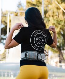 Back view of person in yellow leggings and black t‑shirt with circular logo, strapped into a trapeze safety belt on an outdoor rig, ready to fly.