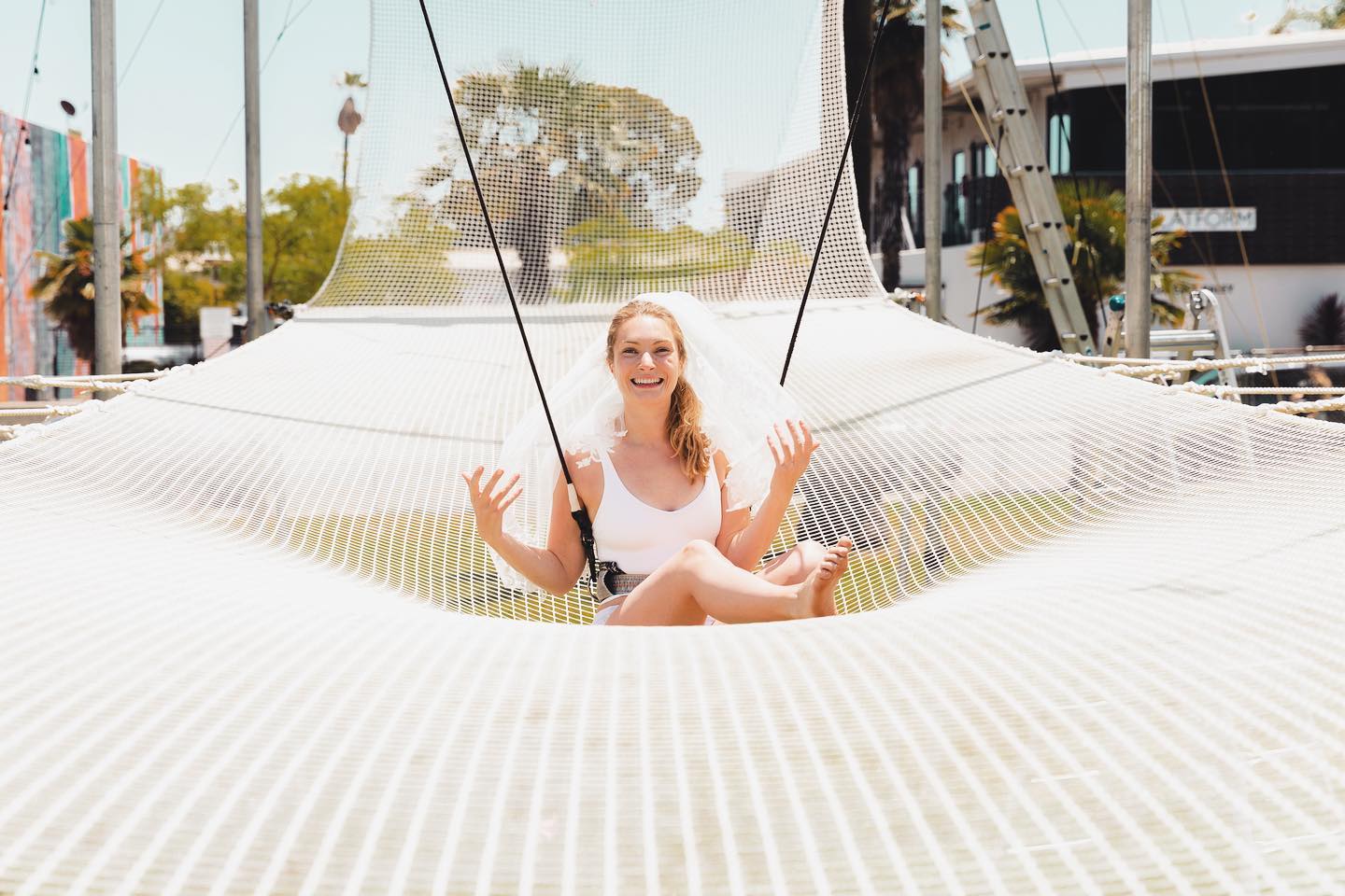 Laughing bride-to-be in a white swimsuit and veil sitting cross-legged on a large suspended white rope-net swing at a sunny outdoor waterfront adventure area