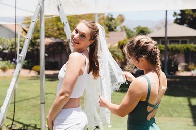 Smiling bride-to-be in a white top with a lace veil while a friend adjusts it outdoors in a sunny backyard wedding-prep scene.