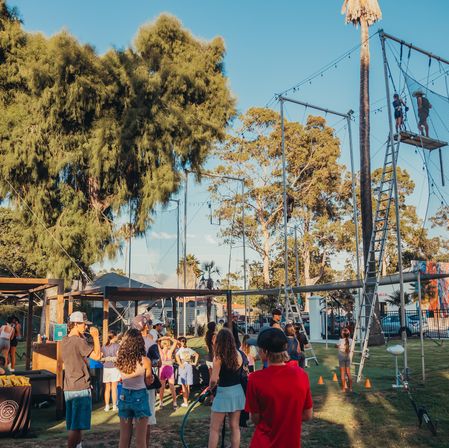 Sunlit community park scene with families gathered around a high-ropes/trapeze course; kids and adults watch participants climb a ladder to a platform framed by tall trees and a palm tree.