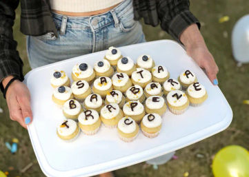 Tray of mini frosted cupcakes topped with blueberries and chocolate letter decorations spelling “Santa Barbara” and additional letters, held outdoors at a casual celebration