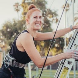 Smiling woman in a safety harness climbing a ladder outdoors in a sunny park with palm trees