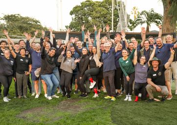 Large diverse group of adults smiling and cheering with hands raised in an outdoor team photo on green grass in a park with palm trees, casual and energetic celebration.