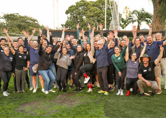 Large diverse group of adults smiling and cheering with hands raised in an outdoor team photo on green grass in a park with palm trees, casual and energetic celebration.