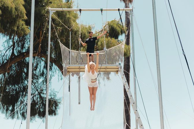 Woman in white hanging from an outdoor trapeze bar over a safety net while an instructor on the platform gives a thumbs-up, framed by palm trees and a clear blue sky — outdoor trapeze lesson vibe.