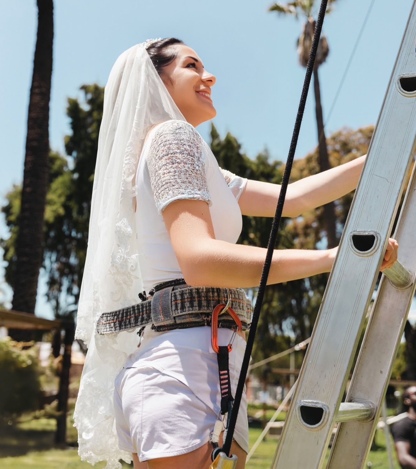 Smiling woman in a wedding veil and lace top wearing a safety harness climbs a ladder in a sunny park with palm trees