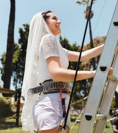 Smiling woman in a wedding veil and lace top wearing a safety harness climbs a ladder in a sunny park with palm trees