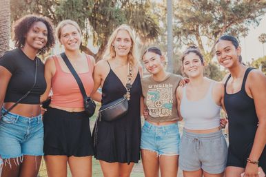 Six smiling young women arm-in-arm in a sunlit park with palm trees, wearing casual summer outfits and crossbody bags