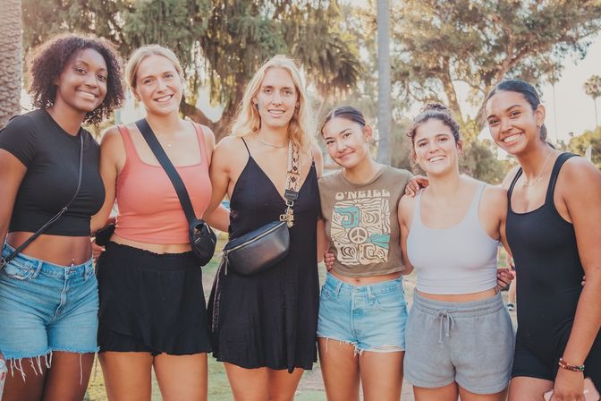 Six smiling young women arm-in-arm in a sunlit park with palm trees, wearing casual summer outfits and crossbody bags
