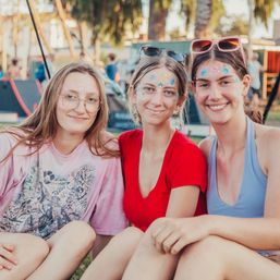 Three smiling young women with face glitter and sunglasses sitting on grass at an outdoor summer festival in a park, enjoying a sunny day.