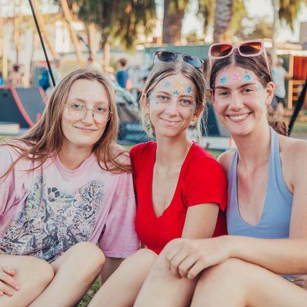 Three smiling young women with face glitter and sunglasses sitting on grass at an outdoor summer festival in a park, enjoying a sunny day.