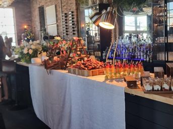 Stylish loft event buffet: white-draped table with carved meat platter, colorful fruit skewers, dessert cups, floral arrangements and mirrored bar backdrop.