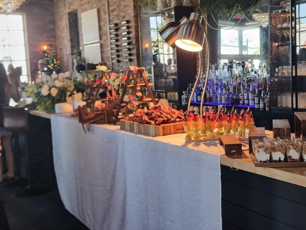Stylish loft event buffet: white-draped table with carved meat platter, colorful fruit skewers, dessert cups, floral arrangements and mirrored bar backdrop.