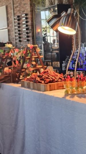 Stylish loft event buffet: white-draped table with carved meat platter, colorful fruit skewers, dessert cups, floral arrangements and mirrored bar backdrop.