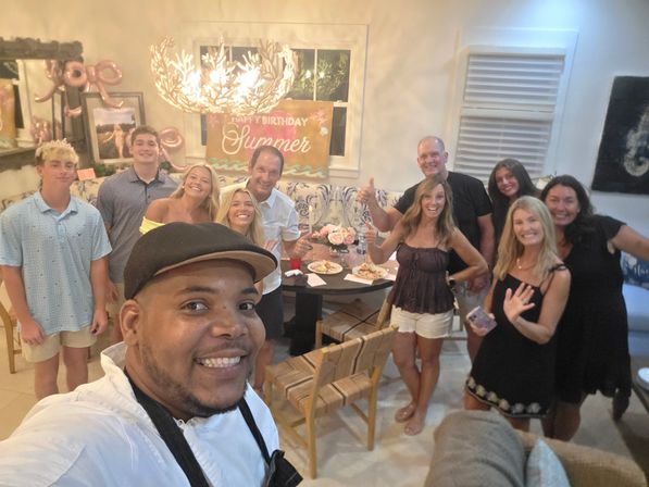 Group selfie at an indoor summer-themed birthday party in a bright dining room, smiling friends and family gathered around a table with plates, flowers and a “Happy Birthday Summer” banner.