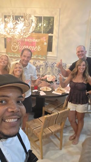 Group selfie at an indoor summer-themed birthday party in a bright dining room, smiling friends and family gathered around a table with plates, flowers and a “Happy Birthday Summer” banner.