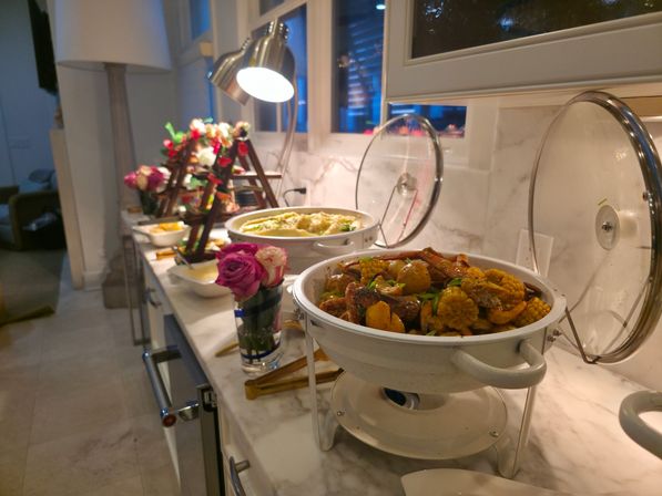 Buffet on a marble kitchen counter with chafing dishes of seasoned seafood boil (corn, potatoes, shrimp), decorative flowers and a task lamp