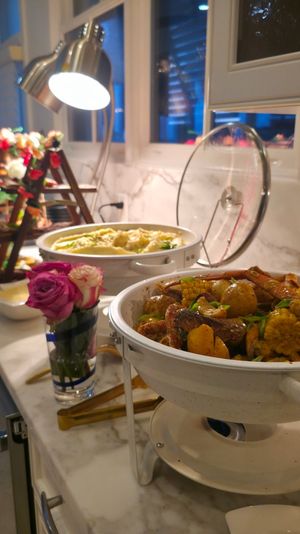 Buffet on a marble kitchen counter with chafing dishes of seasoned seafood boil (corn, potatoes, shrimp), decorative flowers and a task lamp