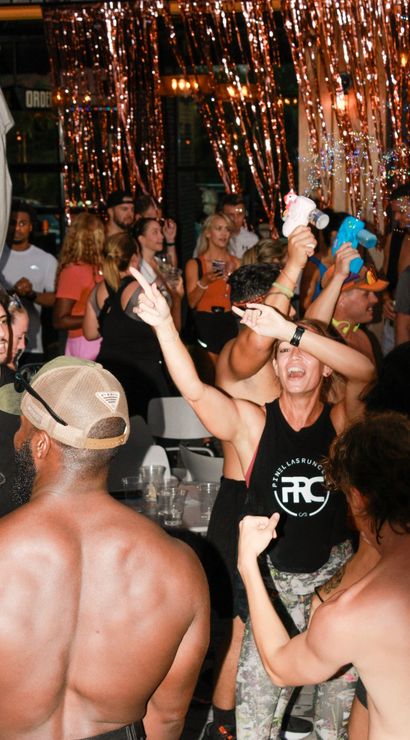 Energetic bar party at night — a crowded group dancing and cheering under metallic streamers and twinkling lights, bubbles and plastic cups, a woman in a black tank top raising her arms while shirtless men dance in the foreground.