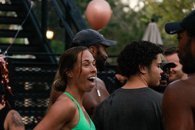 Woman in a green swimsuit top laughing with friends at an outdoor backyard party with balloons and metal stairs in the background.