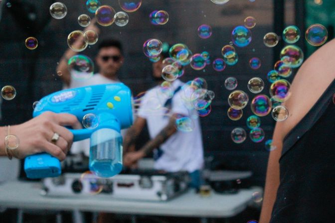Bright blue bubble gun spraying iridescent soap bubbles across an outdoor event, with blurred people and tables in the background.