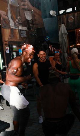 Festive urban outdoor bar patio with a shirtless dancer and smiling friends, soap bubbles drifting past a large beer mural and patio umbrellas.
