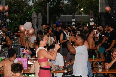 Crowded outdoor patio party at dusk with rose-gold balloons and floating bubbles, people in athletic wear laughing, dancing and taking photos