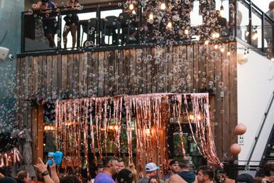 Lively outdoor rooftop patio party with soap bubbles drifting through a crowd, rose-gold fringe curtains, string lights and a wooden facade.