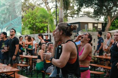 Energetic outdoor beer garden scene with people cheering and laughing around wooden picnic tables, many holding plastic cups of beer beneath palm trees