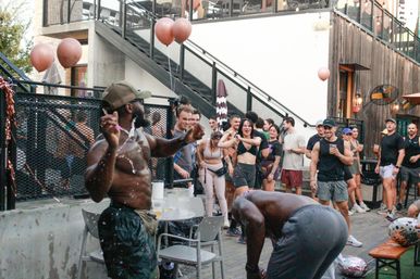 Lively urban outdoor patio party with a shirtless man dancing and splashing water while a crowd in casual summer clothes cheers, holds drinks, and pink balloons float by a metal staircase.