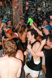 Lively summer party on a bar patio with a crowded group in swimwear dancing and drinking while bubbles from a bubble gun float through the air.