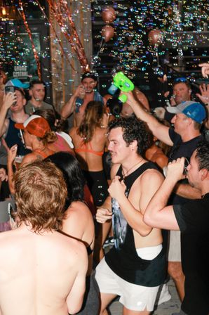 Lively summer party on a bar patio with a crowded group in swimwear dancing and drinking while bubbles from a bubble gun float through the air.