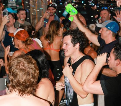 Lively summer party on a bar patio with a crowded group in swimwear dancing and drinking while bubbles from a bubble gun float through the air.