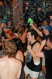 Energetic summer party crowd in swimsuits and tank tops dancing and drinking as colorful soap bubbles float through the air, someone firing a green bubble gun
