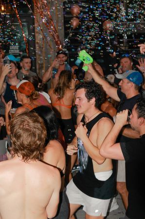Energetic summer party crowd in swimsuits and tank tops dancing and drinking as colorful soap bubbles float through the air, someone firing a green bubble gun