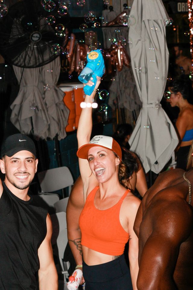 Woman in orange crop top and trucker hat holding a blue bubble gun overhead as iridescent bubbles float around at a lively outdoor nighttime party with friends.